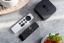 A minimalistic setup with a black streaming device with a logo on top placed on a marble surface. Two sleek, modern remote controls with various buttons are beside it. The arrangement is complemented by decorative plant pots, adding a touch of nature to the scene.