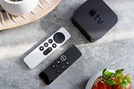 A minimalistic setup with a black streaming device with a logo on top placed on a marble surface. Two sleek, modern remote controls with various buttons are beside it. The arrangement is complemented by decorative plant pots, adding a touch of nature to the scene.