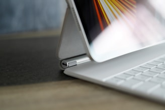A close-up view of a tablet propped up by a folio case on a keyboard cover. The screen displays abstract lines in bright colors, and the focus is on the hinge of the case.