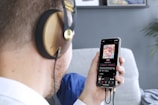 A person wearing large headphones is holding a smartphone in one hand. On the smartphone screen, a colorful album cover with floral patterns is visible, along with music playing details. The scene includes a blurred background with a grey wall and some framed art.