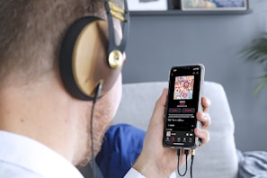A person wearing large headphones is holding a smartphone in one hand. On the smartphone screen, a colorful album cover with floral patterns is visible, along with music playing details. The scene includes a blurred background with a grey wall and some framed art.