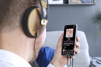 A person wearing large headphones is holding a smartphone in one hand. On the smartphone screen, a colorful album cover with floral patterns is visible, along with music playing details. The scene includes a blurred background with a grey wall and some framed art.