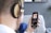 A person wearing large headphones is holding a smartphone in one hand. On the smartphone screen, a colorful album cover with floral patterns is visible, along with music playing details. The scene includes a blurred background with a grey wall and some framed art.