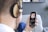A person wearing large headphones is holding a smartphone in one hand. On the smartphone screen, a colorful album cover with floral patterns is visible, along with music playing details. The scene includes a blurred background with a grey wall and some framed art.