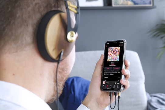 A person wearing large headphones is holding a smartphone in one hand. On the smartphone screen, a colorful album cover with floral patterns is visible, along with music playing details. The scene includes a blurred background with a grey wall and some framed art.
