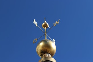 A golden weather vane is mounted on top of a spherical ornament against a clear blue sky. The vane indicates directions such as north and west with metal letters attached to it.