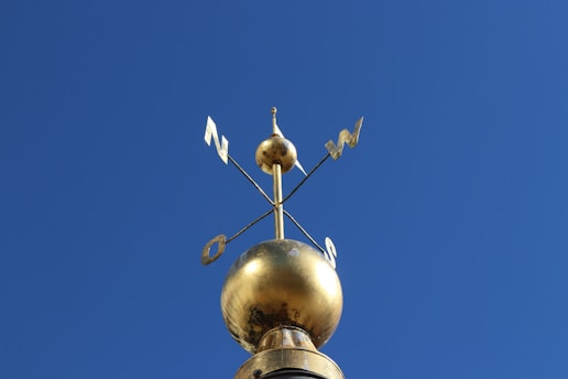 A golden weather vane is mounted on top of a spherical ornament against a clear blue sky. The vane indicates directions such as north and west with metal letters attached to it.