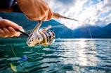 A person is holding a fishing rod, casting it over a large body of water surrounded by mountains. The sun is shining brightly, reflecting off the surface of the water. The scene is serene, with a backdrop of partly cloudy sky and distant mountains.