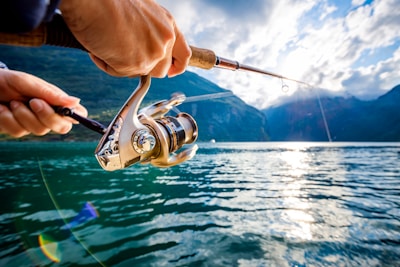 A person is holding a fishing rod, casting it over a large body of water surrounded by mountains. The sun is shining brightly, reflecting off the surface of the water. The scene is serene, with a backdrop of partly cloudy sky and distant mountains.