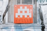 A pixelated, gray alien figure is prominently displayed on a red square panel with graffiti-like markings below it. The panel is mounted on a wall with a metal mesh background, surrounded by posters and faded graffiti.