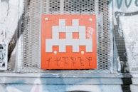 A pixelated, gray alien figure is prominently displayed on a red square panel with graffiti-like markings below it. The panel is mounted on a wall with a metal mesh background, surrounded by posters and faded graffiti.