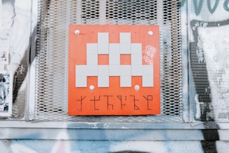 A pixelated, gray alien figure is prominently displayed on a red square panel with graffiti-like markings below it. The panel is mounted on a wall with a metal mesh background, surrounded by posters and faded graffiti.
