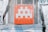 A pixelated, gray alien figure is prominently displayed on a red square panel with graffiti-like markings below it. The panel is mounted on a wall with a metal mesh background, surrounded by posters and faded graffiti.