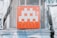 A pixelated, gray alien figure is prominently displayed on a red square panel with graffiti-like markings below it. The panel is mounted on a wall with a metal mesh background, surrounded by posters and faded graffiti.