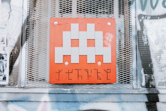 A pixelated, gray alien figure is prominently displayed on a red square panel with graffiti-like markings below it. The panel is mounted on a wall with a metal mesh background, surrounded by posters and faded graffiti.