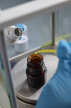 A person wearing blue gloves is using a pipette to transfer liquid into a small brown glass bottle. The setup appears to be in a laboratory with other equipment and bottles in the background.