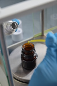 A person wearing blue gloves is using a pipette to transfer liquid into a small brown glass bottle. The setup appears to be in a laboratory with other equipment and bottles in the background.
