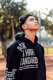 A young man gazes upward with a reflective expression. He is wearing a black hoodie with bold white text, set against a blurred background of trees and sunlight filtering through the leaves.