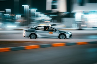 A taxi car is driving swiftly along a city street at night, with lights blurred in the background indicating motion and speed.