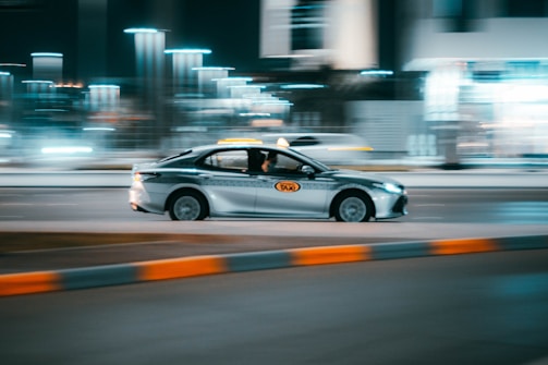 A taxi car is driving swiftly along a city street at night, with lights blurred in the background indicating motion and speed.