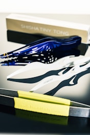 A set of stylish and contemporary tongs, with distinct designs, rest on a polished black surface. One tong is metallic with a yellow rubber grip, another features a sleek white finish, and a third one showcases a glossy blue coloration. In the background, there is a partially visible box labeled 'Shisha Tiny Tong'. The reflective surface below adds an abstract mirroring effect to the overall scene.