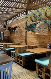 A rustic-themed seating area features wooden tables and booths with light blue cushions. Overhead, woven rattan light fixtures hang from a slatted wooden ceiling, casting soft shadows. Surfboards with floral designs and a brand logo are mounted on the wooden wall, adding a coastal ambiance. The floors are made of polished wood, enhancing the natural aesthetic.