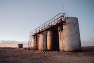 A group of large, cylindrical storage tanks is located on a barren, gravel-covered landscape. The tanks are connected by a metal catwalk with stairs leading to the top. The sky in the background is clear with subtle hues of a sunset or sunrise, casting a soft pink and orange glow.