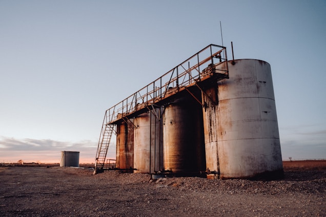 A group of large, cylindrical storage tanks is located on a barren, gravel-covered landscape. The tanks are connected by a metal catwalk with stairs leading to the top. The sky in the background is clear with subtle hues of a sunset or sunrise, casting a soft pink and orange glow.