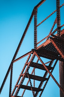 A metal structure framing the sky with stair-like features, characterized by industrial design with holes in the metal beams creating a patterned texture. The structure appears sturdy and functional, intersecting at various angles.