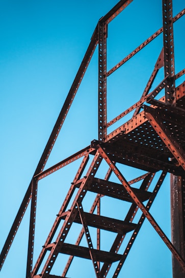 A metal structure framing the sky with stair-like features, characterized by industrial design with holes in the metal beams creating a patterned texture. The structure appears sturdy and functional, intersecting at various angles.