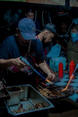 Several people are gathered around a cooking station at what appears to be a street food market. A man in the foreground is using a blowtorch to grill skewered food on a metal rack. There are containers filled with food items and condiments like ketchup bottles on a checkered tablecloth. The lighting is dim, giving the scene an intimate, bustling atmosphere.