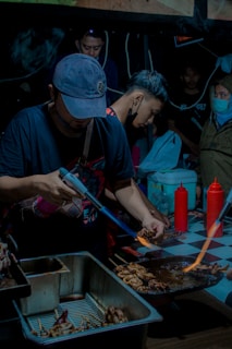 Several people are gathered around a cooking station at what appears to be a street food market. A man in the foreground is using a blowtorch to grill skewered food on a metal rack. There are containers filled with food items and condiments like ketchup bottles on a checkered tablecloth. The lighting is dim, giving the scene an intimate, bustling atmosphere.