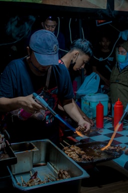Several people are gathered around a cooking station at what appears to be a street food market. A man in the foreground is using a blowtorch to grill skewered food on a metal rack. There are containers filled with food items and condiments like ketchup bottles on a checkered tablecloth. The lighting is dim, giving the scene an intimate, bustling atmosphere.