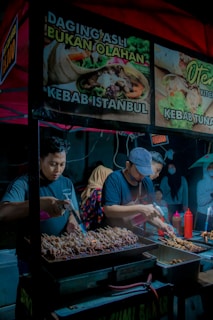 Two people are preparing skewered food on a grill under a brightly colored canopy. The surrounding area is a vibrant street market setting. Signage above advertises various kebab options.