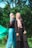 Two women wearing hijabs stand outdoors surrounded by lush greenery. One is in blue pants holding a small decoration, and the other is holding a smartphone. The background includes trees and a green tent.