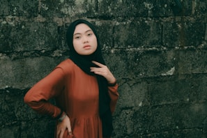 A confident young woman wearing a dusty rose hijab, smiling softly against a cream-colored wall.