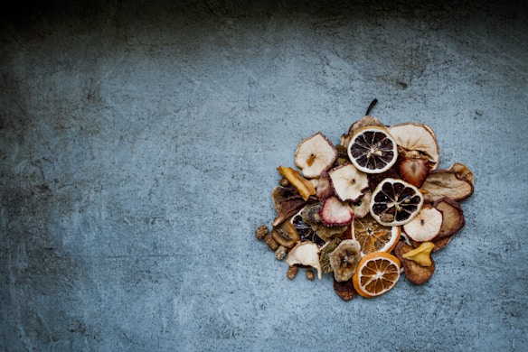 A collection of various dried fruit slices, including oranges, apples, and lemons, arranged on a textured, blue-toned background. The fruits display a range of colors and textures, creating a contrast against the muted surface they rest on.
