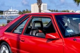 A vibrant red sports car parked in an outdoor setting with a glimpse of trees, buildings, and a body of water in the background. The car's design features include large side windows, a visible seat with red accents, and a sticker that reads 'Let me guess... License & Registration'.
