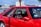 A vibrant red sports car parked in an outdoor setting with a glimpse of trees, buildings, and a body of water in the background. The car's design features include large side windows, a visible seat with red accents, and a sticker that reads 'Let me guess... License & Registration'.