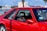 A vibrant red sports car parked in an outdoor setting with a glimpse of trees, buildings, and a body of water in the background. The car's design features include large side windows, a visible seat with red accents, and a sticker that reads 'Let me guess... License & Registration'.