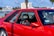 A vibrant red sports car parked in an outdoor setting with a glimpse of trees, buildings, and a body of water in the background. The car's design features include large side windows, a visible seat with red accents, and a sticker that reads 'Let me guess... License & Registration'.