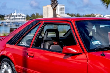 A vibrant red sports car parked in an outdoor setting with a glimpse of trees, buildings, and a body of water in the background. The car's design features include large side windows, a visible seat with red accents, and a sticker that reads 'Let me guess... License & Registration'.