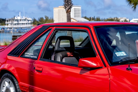 A vibrant red sports car parked in an outdoor setting with a glimpse of trees, buildings, and a body of water in the background. The car's design features include large side windows, a visible seat with red accents, and a sticker that reads 'Let me guess... License & Registration'.