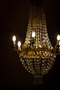 An ornate crystal chandelier adorned with gold metalwork and illuminated candle-style bulbs, casting a warm light. The chandelier features multiple crystal strands, cascading down in a conical shape, creating a luxurious and opulent atmosphere. The background appears dark, accentuating the chandelier's glow.