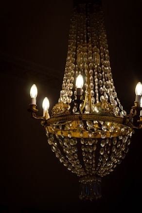 An ornate crystal chandelier adorned with gold metalwork and illuminated candle-style bulbs, casting a warm light. The chandelier features multiple crystal strands, cascading down in a conical shape, creating a luxurious and opulent atmosphere. The background appears dark, accentuating the chandelier's glow.