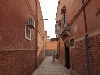 A narrow alleyway flanked by terracotta-colored buildings, featuring an air conditioning unit on the left and various signs indicating nearby hotels. The pathway is paved with cobblestones, and a lone figure is seen walking towards the end of the alley.