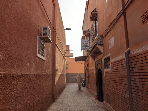 A narrow alleyway flanked by terracotta-colored buildings, featuring an air conditioning unit on the left and various signs indicating nearby hotels. The pathway is paved with cobblestones, and a lone figure is seen walking towards the end of the alley.