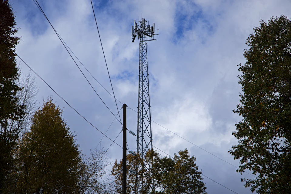 The image depicts a tall, gray metal cell tower standing against a clear blue sky with fluffy white clouds. The tower is situated on the right side of the frame and reaches up into the sky. In the background, there are trees visible, adding depth to the scene.
