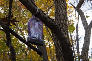 A plastic or resin tombstone decoration is hanging by a string from a branch, set against a backdrop of autumn trees with leaves in shades of green, yellow, and brown.