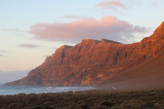 An expansive view of a dramatic coastline with towering cliffs bathed in warm sunlight. The ocean meets the rugged terrain, and the sky is scattered with soft, pinkish clouds.